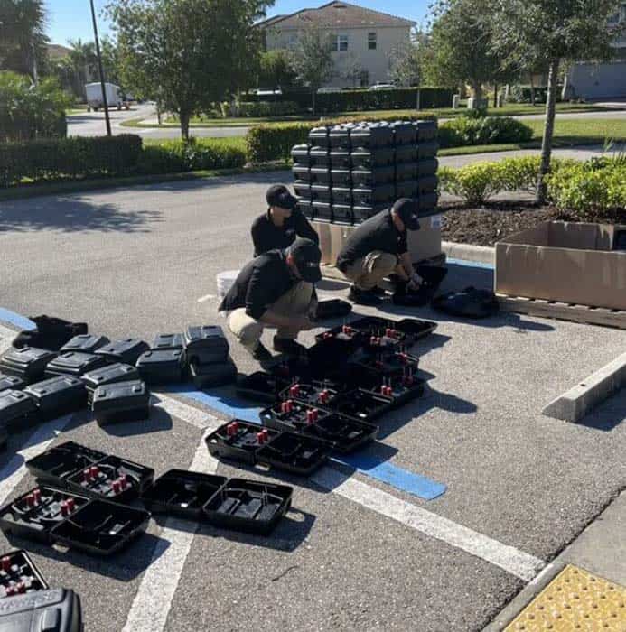 Three people in Stat Pest Control uniforms are organizing or assembling black hard cases with red interiors in a parking lot. Stacks of closed cases and a cardboard box are nearby, with trees and buildings in the background.