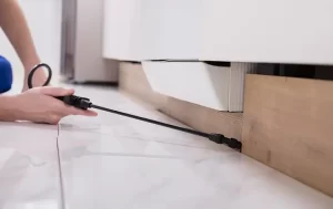 A person from Stat Pest Control uses a spray wand to treat the area under a kitchen cabinet along the baseboard. The tiled floor and a partially open drawer are also visible.