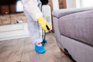 A technician from Stat Pest Control, dressed in protective clothing with yellow gloves and blue shoe covers, sprays near the base of a gray sofa in a living room, likely performing pest control or cleaning.