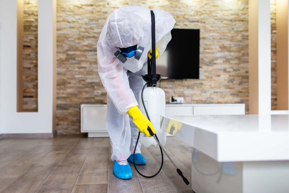 A Stat Pest Control technician in protective gear, including gloves, mask, and coveralls, uses a sprayer to disinfect or treat the surface of a white table in a modern indoor setting with wooden flooring.
