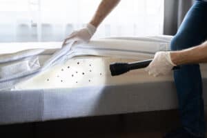 A Stat Pest Control expert wearing gloves lifts a mattress cover and shines a flashlight on the mattress, revealing multiple black spots on the surface, possibly indicating a pest infestation.